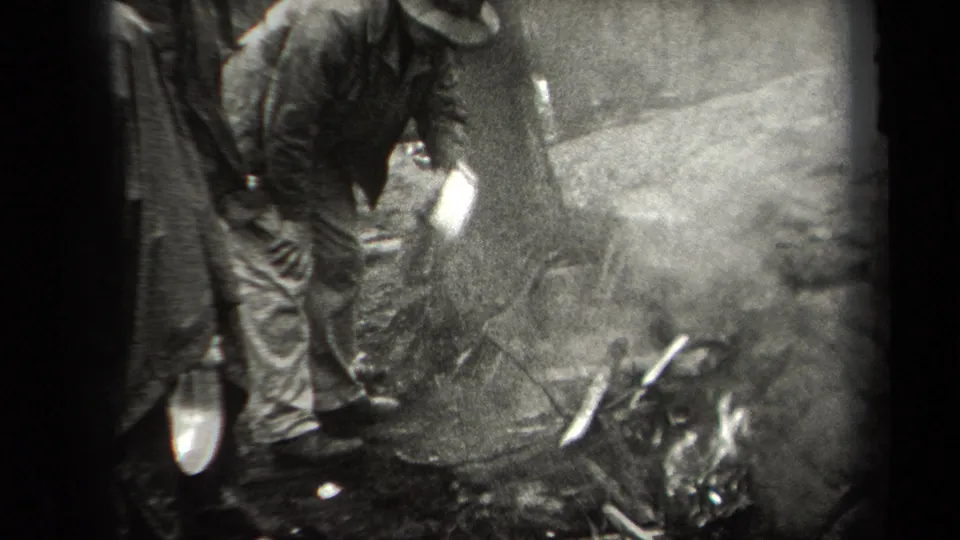 1938 New York home-movie frame with a person tending a smoky outdoor fire pit beside a hanging chicken and rough ground.