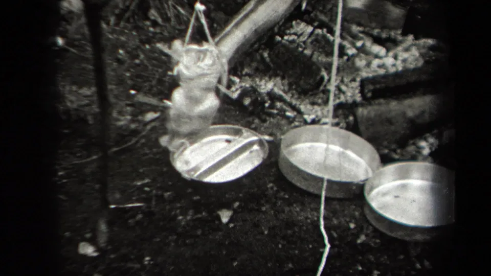 1938 New York home-movie frame of a chicken suspended by string near metal pans, burned wood, and a rough outdoor hearth.