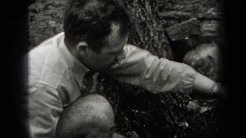 An adult man in a light shirt and tie crouches near the wooded cook-fire area in the supplied 1938 New York home-movie footage.