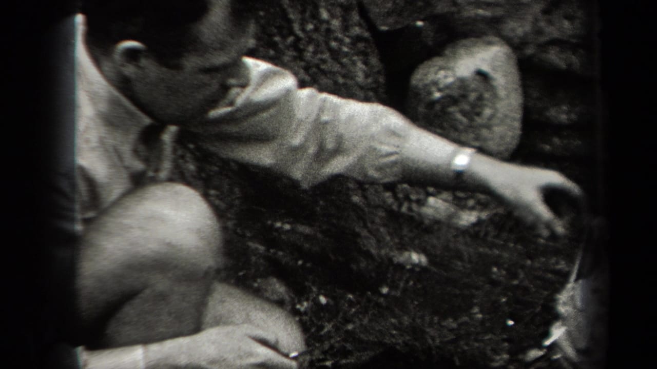 In 1938 New York home-movie footage, a man in a light shirt crouches in woods near a rough cooking setup.
