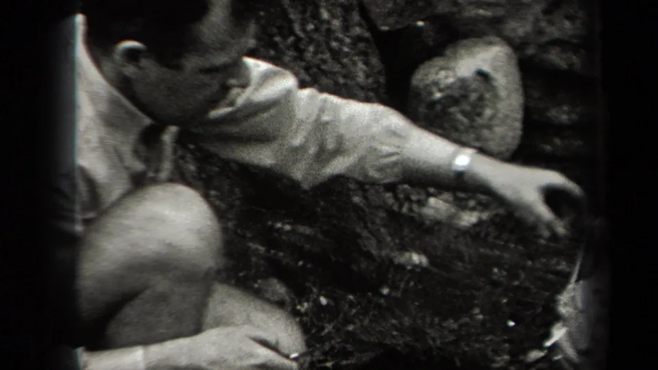 In 1938 New York home-movie footage, a man in a light shirt crouches in woods near a rough cooking setup.