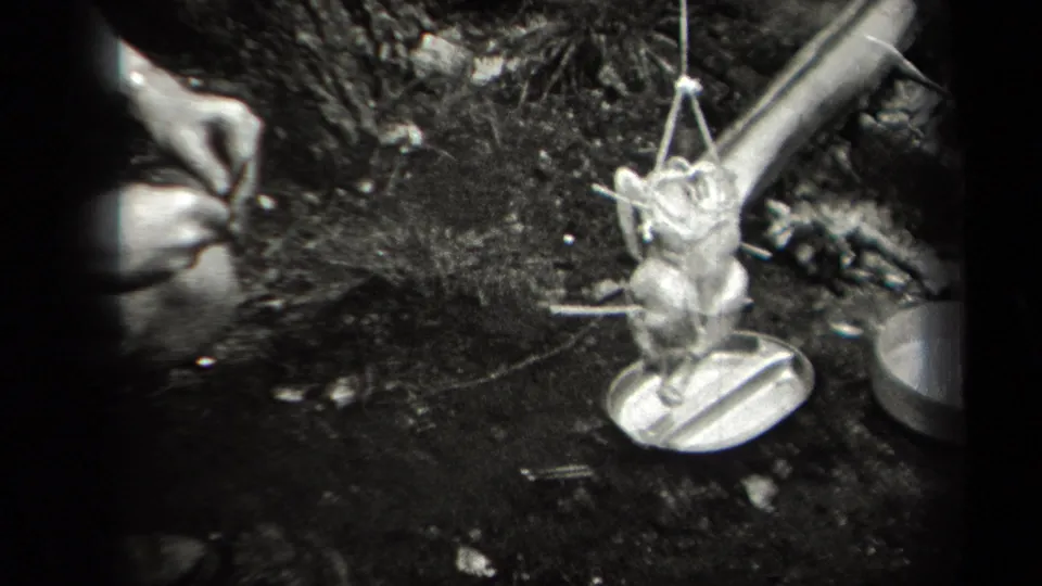 A whole bird is suspended from a makeshift support over a campfire area in wooded 1938 New York footage.