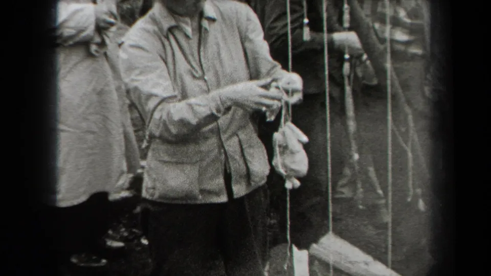 The home-movie frame shows a person adjusting a chicken suspended by string, with other adults gathered nearby in New York, 1938.