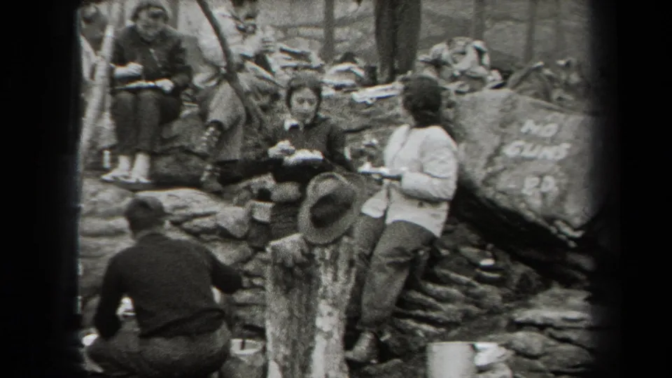 1938 New York home-movie frame showing campers eating near rocks, trees, smoke, and a large stone marked NO GUNS.