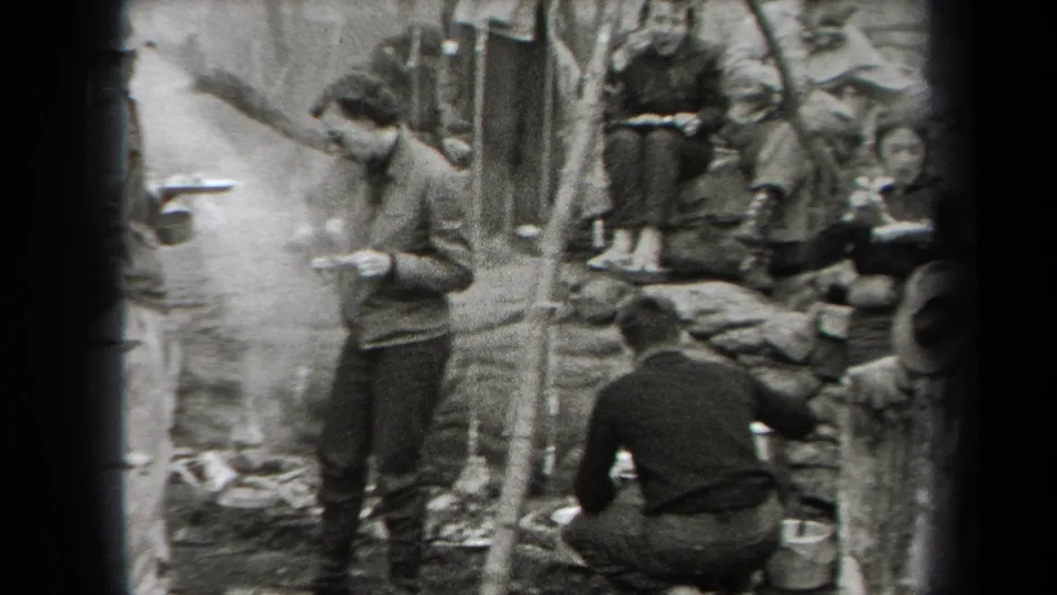 1938 New York campsite frame with a standing man holding food as seated campers gather beside the smoky fire area.