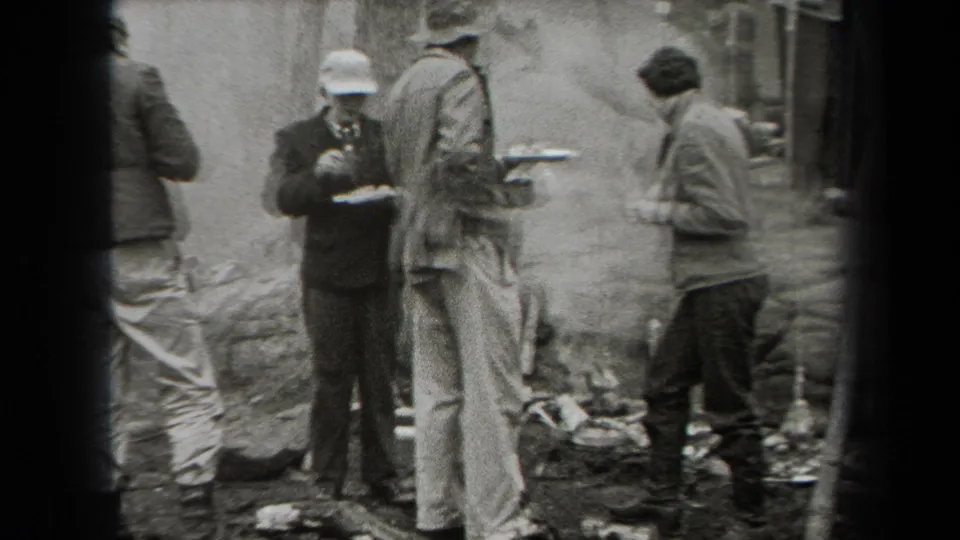 1938 New York outdoor camp frame showing men standing by a smoky fire pit, with plates, boots, rocks, and trees visible.