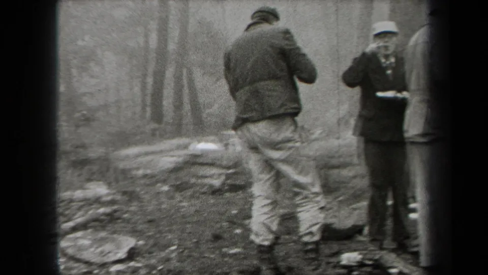 1938 New York campsite frame with two men at the edge of a smoky fire area amid rocks, trees, boots, and outdoor clothing.