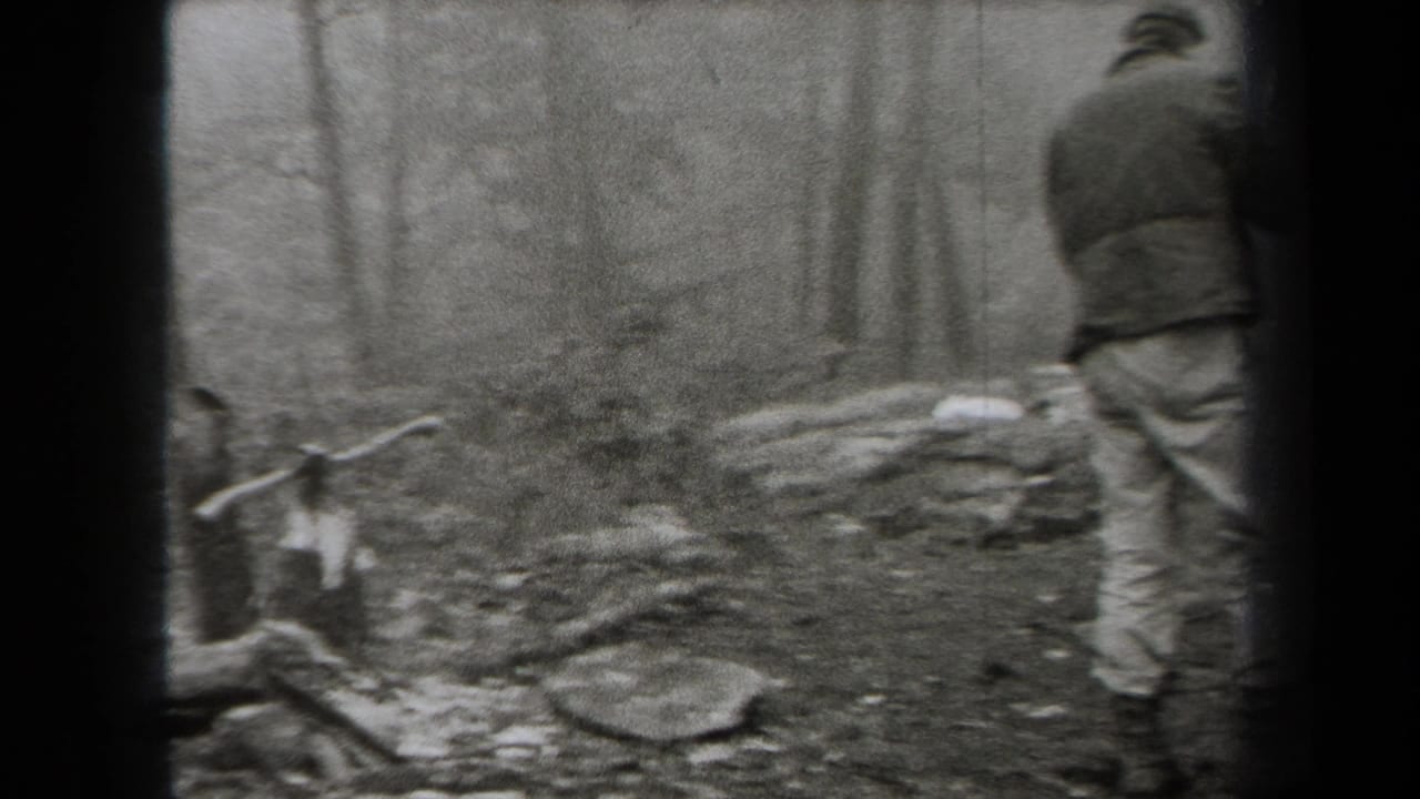 1938 New York home-movie frame showing hikers in coats moving through a forested area with rocks, bare trees, and fallen branches.