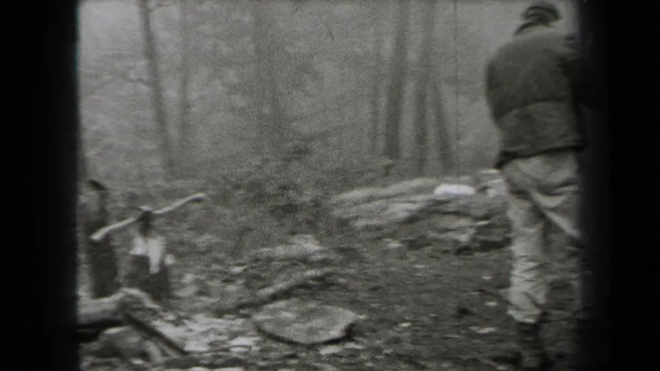 1938 New York home-movie frame showing hikers in coats moving through a forested area with rocks, bare trees, and fallen branches.