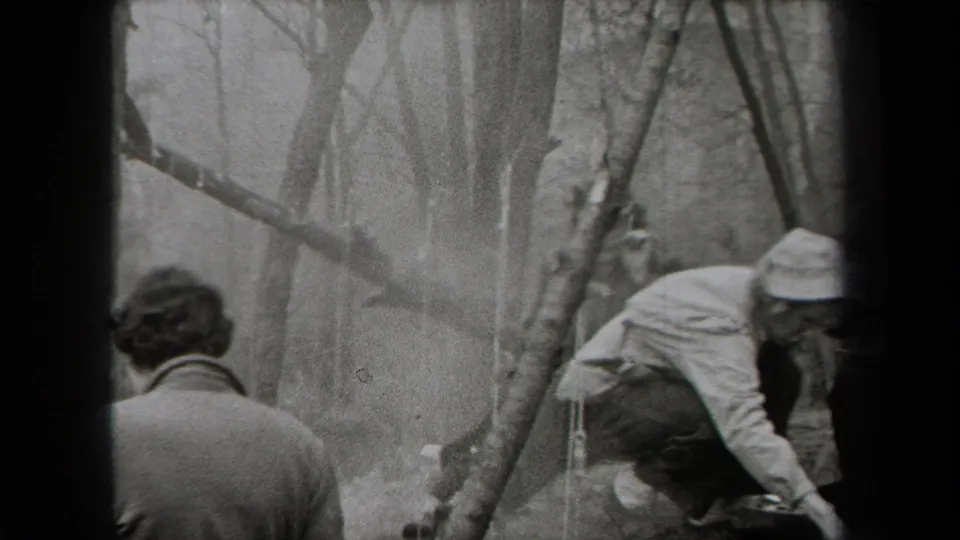In a New York woodland meal stop, a hiker bends near smoke and cooking gear while another figure is seen from behind.