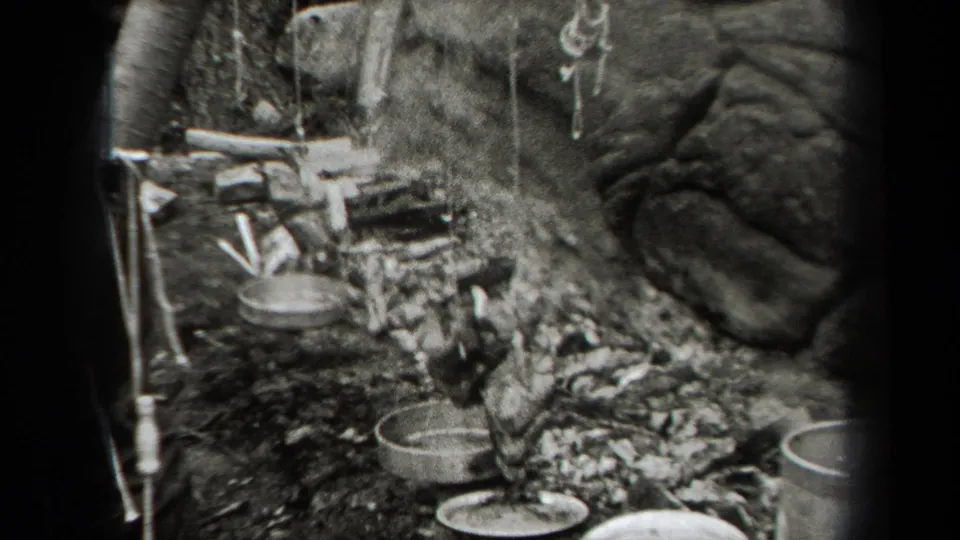 Close view of a woodland cooking setup with pots, cups, mess kits, firewood, and a small campfire beside a tree during the 1938 hiking stop.