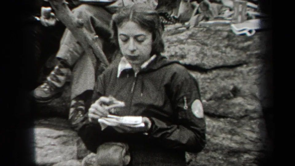 In 1938 New York home-movie footage, a young woman in a dark jacket eats from a small plate near rocks and other seated adults.