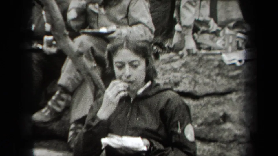 A young woman in a dark jacket eats outdoors from a plate in the 1938 New York home-movie scene, with rocks and seated companions behind her.
