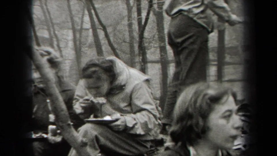 Adults in jackets sit among rocks and bare trees, eating from plates in black-and-white 1938 New York home-movie footage.
