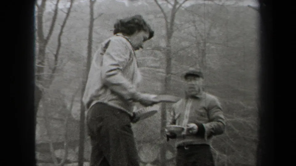 A woman holding a plate stands near a seated man in a cap, with bare trees behind them in the 1938 New York outdoor meal footage.