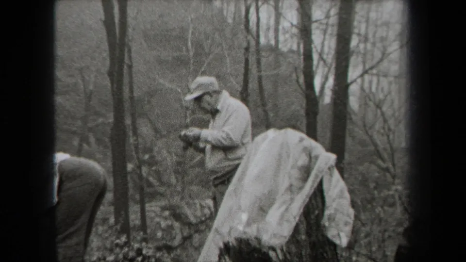 Black-and-white 1938 New York frame with a capped man standing in woodland beside a covered object or cloth.