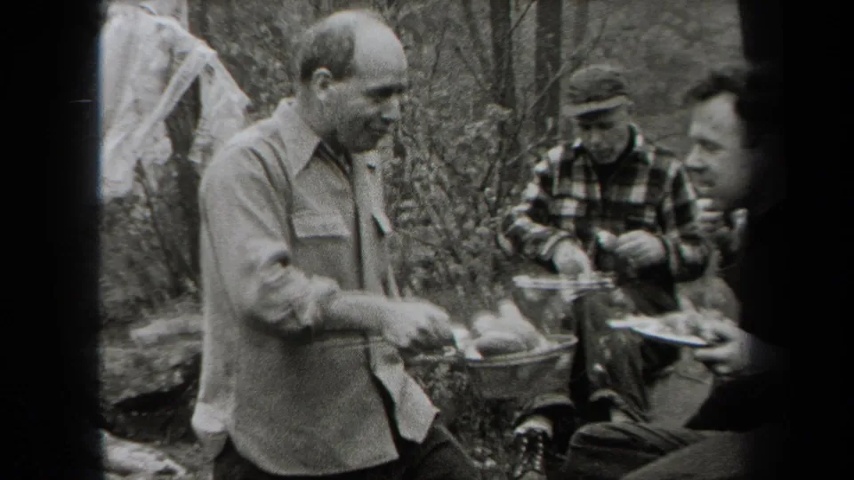 Black-and-white 1938 New York home-movie frame showing men with plates during an outdoor meal in a wooded setting.