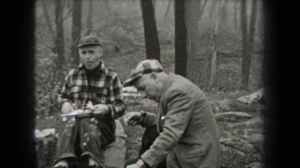 Men in caps and plaid outdoor clothing eat beside rocks in a wooded New York mountain area.