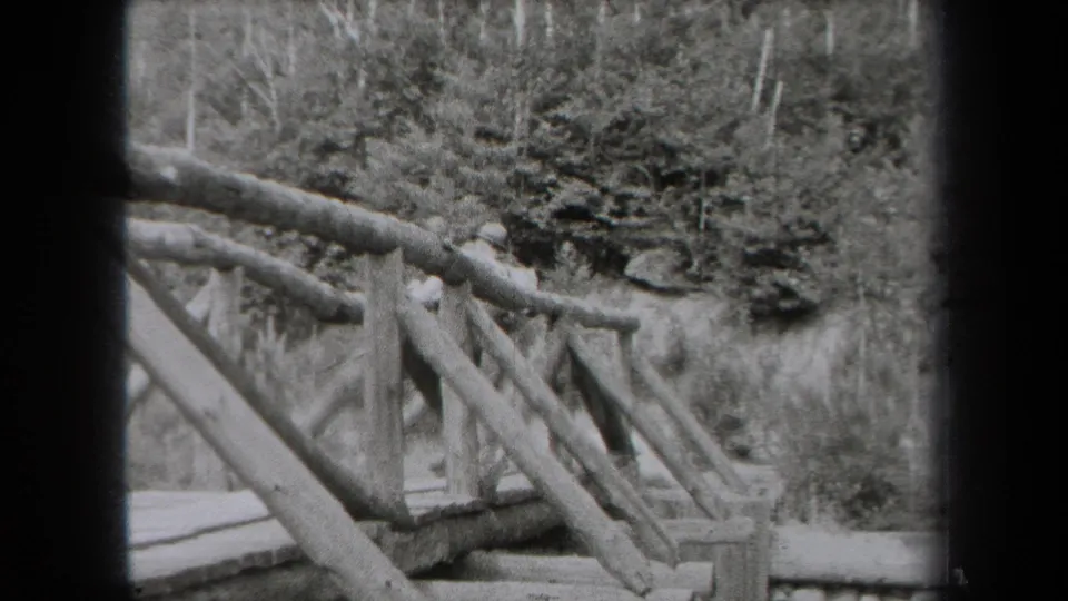 Black-and-white New York home-movie view of a rough wooden bridge rail above trees, rocks, and a stream area.