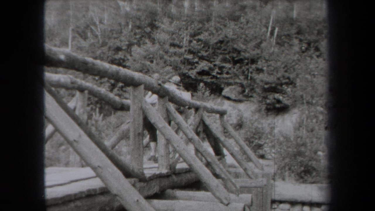 1938 New York 8mm frame showing a rustic wooden bridge railing near trees, rocks, and a calm lakeside setting.