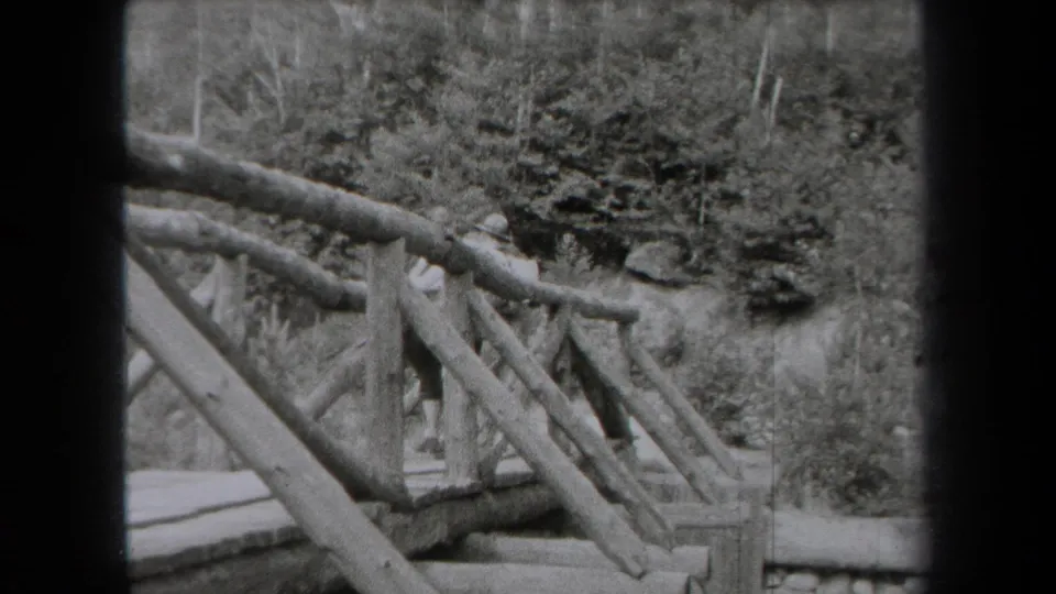 1938 New York 8mm frame showing a rustic wooden bridge railing near trees, rocks, and a calm lakeside setting.