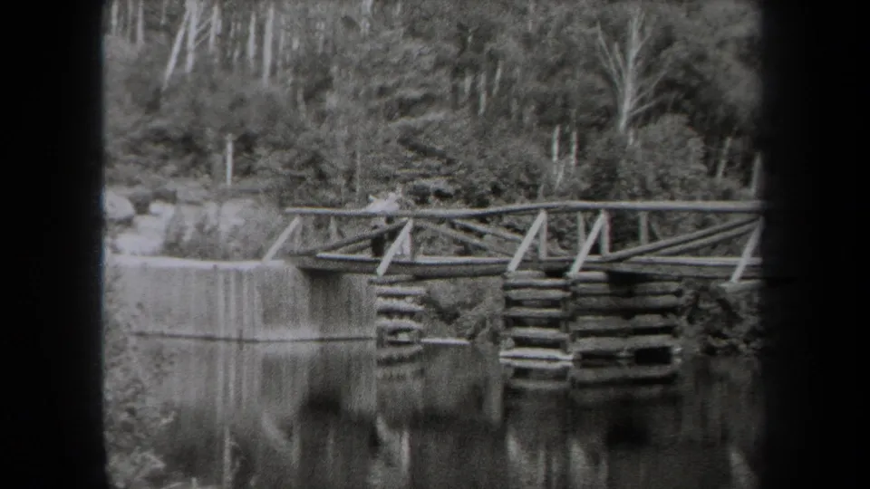 A rustic footbridge spans still water in a wooded New York park, with reflections and dense trees visible in the 1938 home-movie frame.