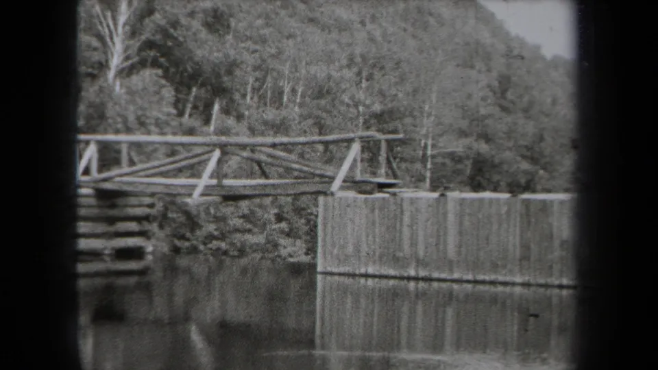 1938 New York park footage shows a rustic bridge over water with a low lakeside wall and wooded hillside behind it.