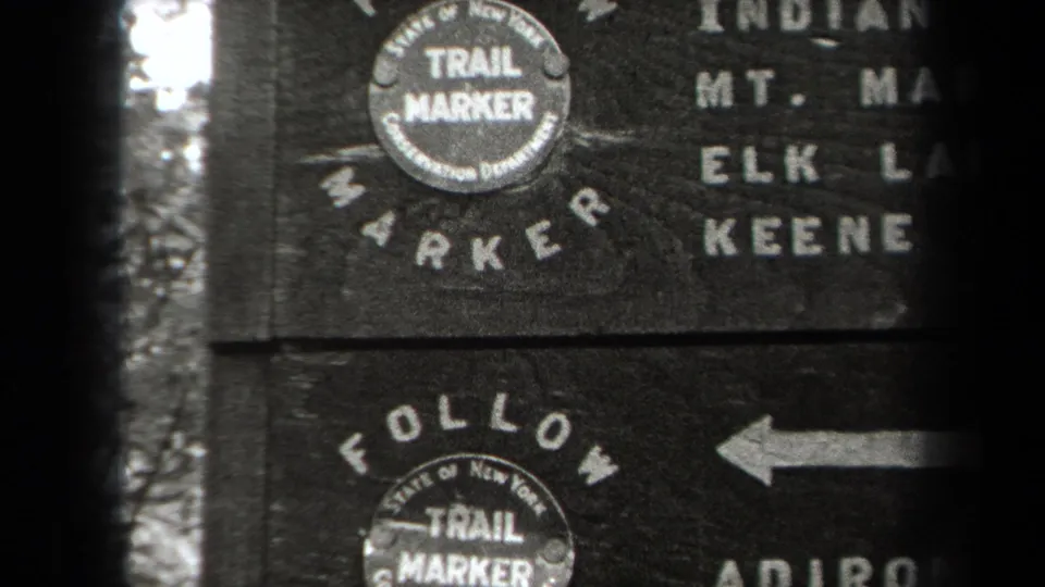 Close-up of a State of New York trail marker sign, with partial destination names and an arrow visible in the 1938 8mm frame.