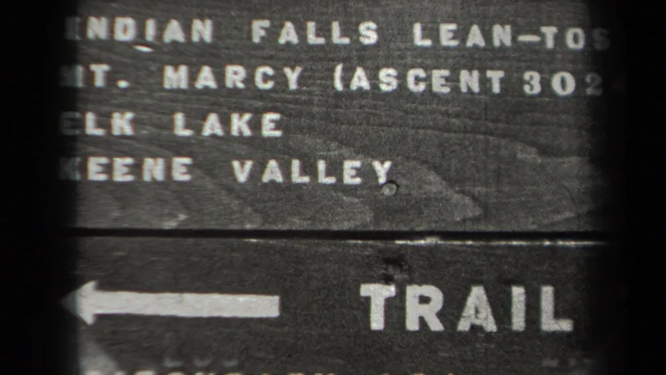 Close 8mm view of the New York trail marker text, with Adirondack destinations painted across dark wooden boards.