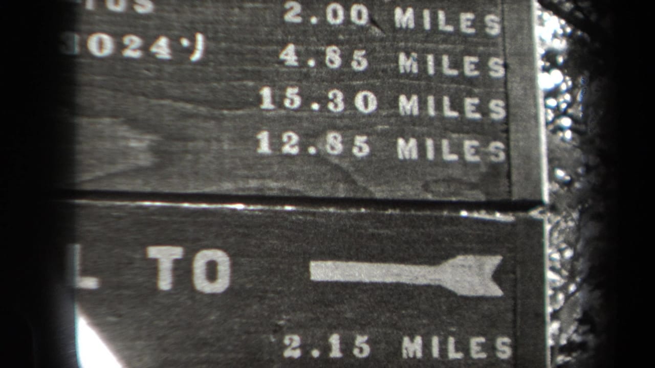 1938 New York 8mm frame showing a weathered wooden trail marker with white mileage figures and directional arrows in a wooded setting.
