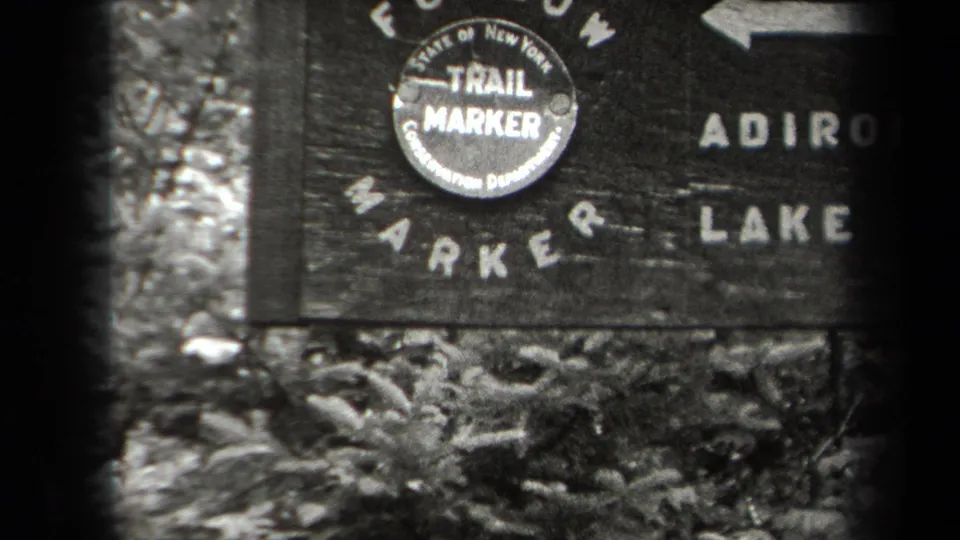 Close 1938 New York frame of a wooden Adirondack-area sign with a round Conservation Department trail marker emblem and arrow lettering.