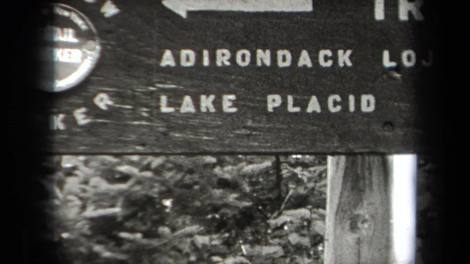 1938 New York 8mm frame showing partial Adirondack and Lake Placid lettering on a wooden trail sign in the woods.