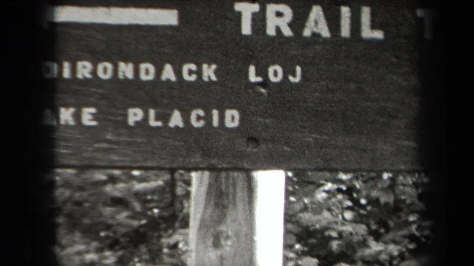 Cropped 1938 New York frame of a wooden trail sign showing trail text, partial Adirondack lettering, Lake Placid, and a support post.