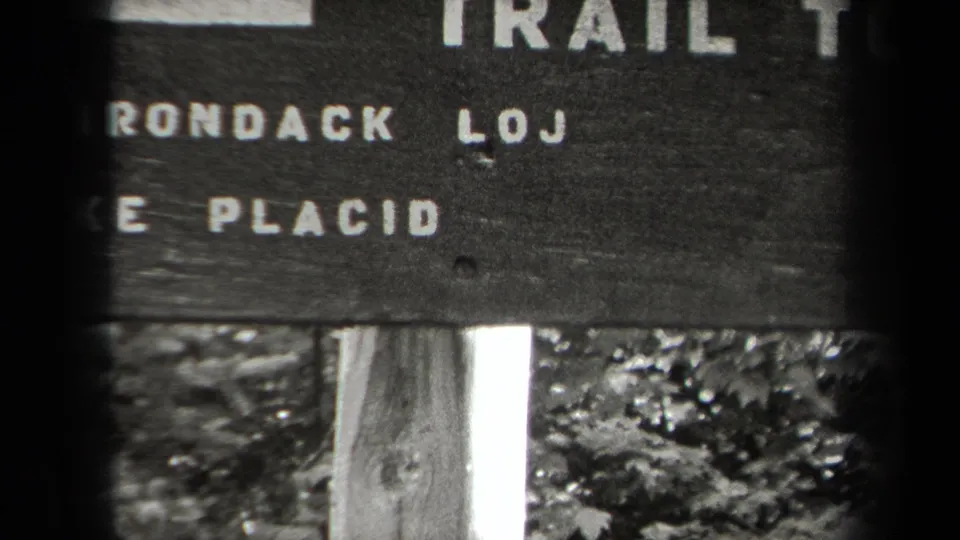 1938 Adirondacks home-movie frame showing a weathered wooden trail sign with partial place names and mileage text.