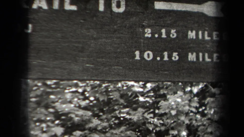 Black-and-white 8mm frame of the Adirondack trail sign, emphasizing mileage numbers and surrounding woodland plants.