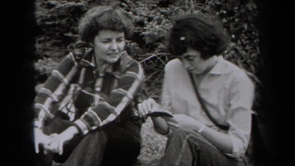 Two women sit close together on a New York hillside in 1938 home-movie footage, appearing relaxed beside shrubs and rocks.