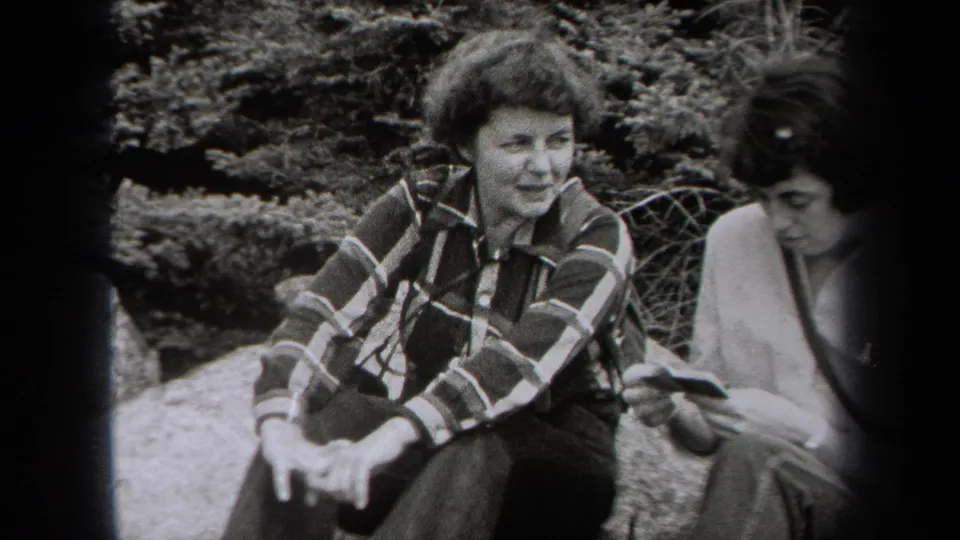 In metadata-supported 1938 New York footage, two women sit outdoors near shrubs, with one smiling in a plaid shirt and the other looking down.