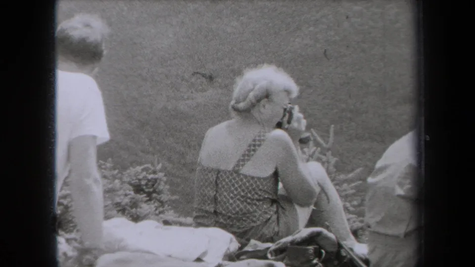 A woman in a patterned sleeveless top sits on grass in the 1938 New York home movie, holding a small camera or object near her face.