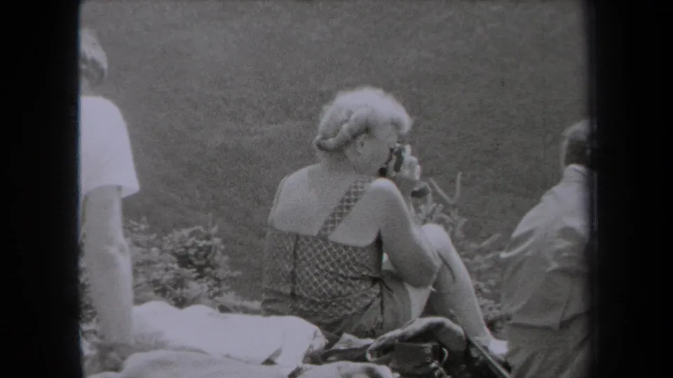The 1938 New York home-movie frame shows a woman seated on grass with a small camera or object, with partial figures and a blanket nearby.