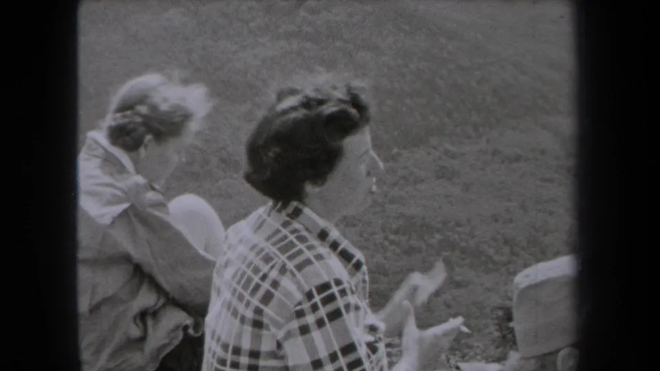 A late-1930s New York home-movie frame shows women seated on grass, including a plaid-shirted figure seen from behind and a companion nearby.