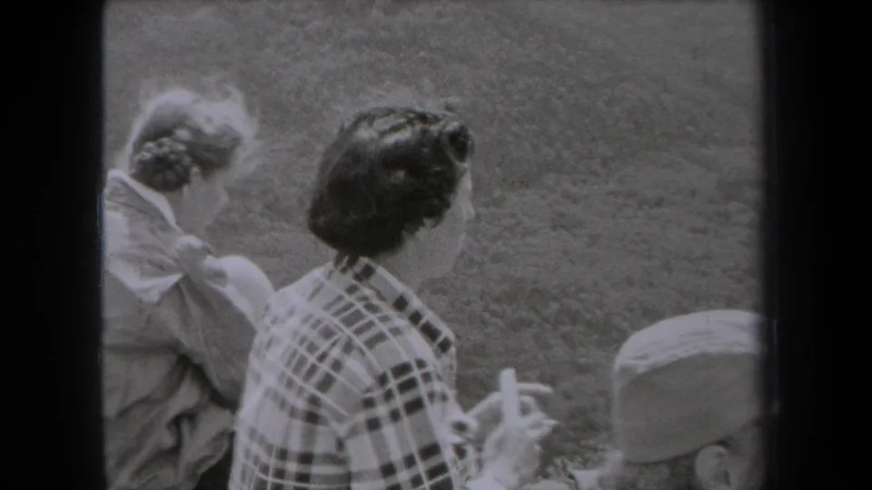 1938 New York home-movie frame of a woman in a plaid shirt holding a white comb while seated outdoors beside another person.
