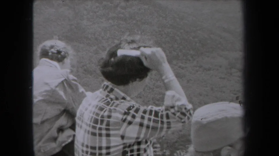 1938 New York frame showing a seated woman combing her dark hair with a white comb in a grassy outdoor setting.