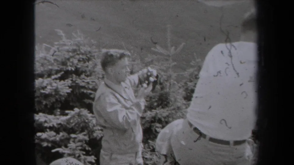 1938 New York outdoor frame with a man holding a camera near bushes as seated companions remain partly visible.