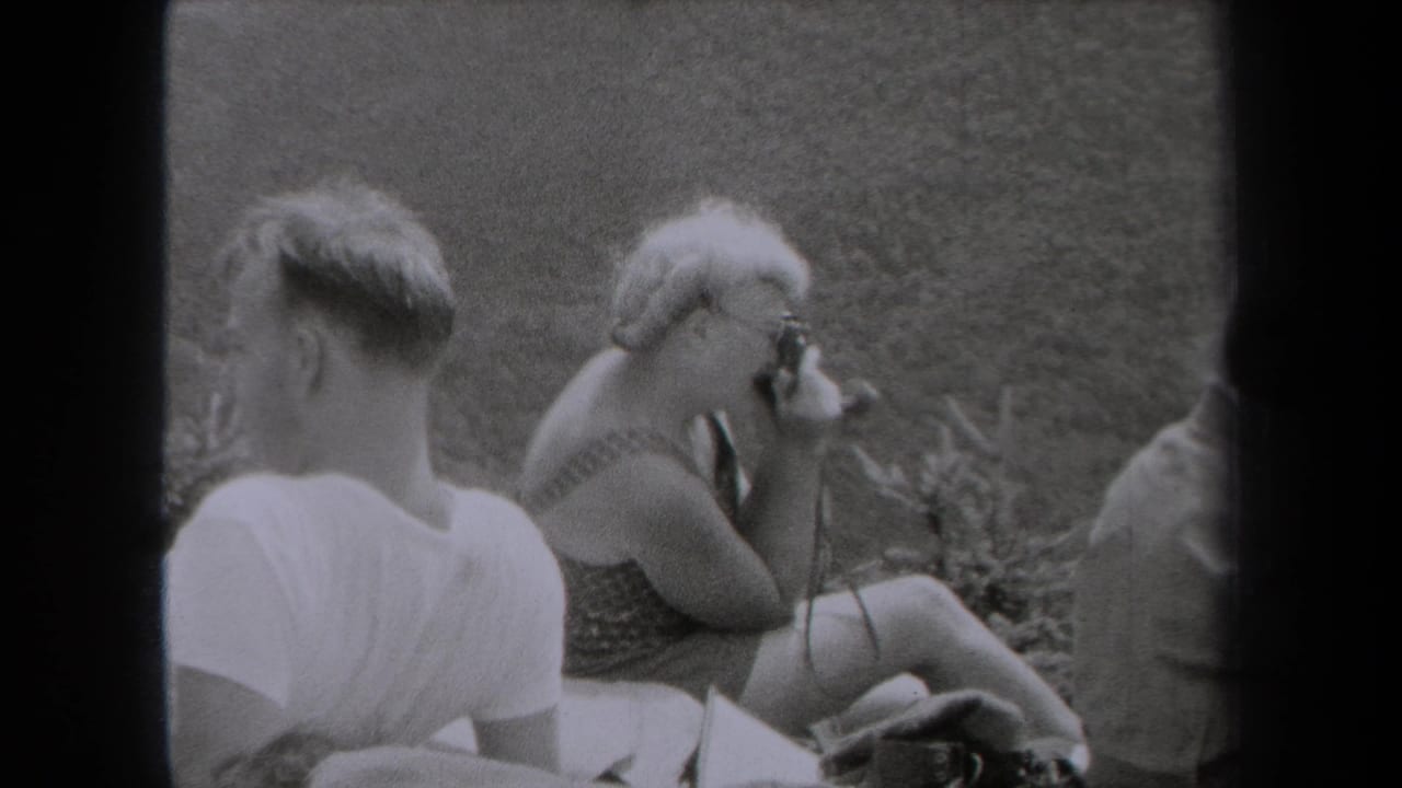 A man in a white T-shirt sits near a woman holding a camera on a grassy hillside.