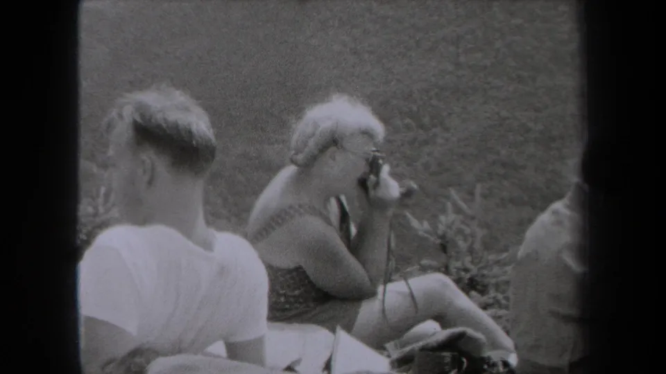 1938 New York frame with a seated man in a white T-shirt and a woman using a camera beside others on grass.