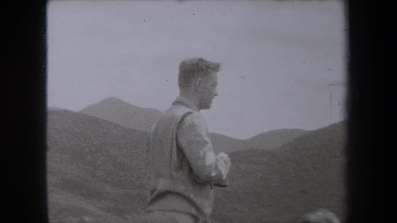 Man in a vest stands before mountain slopes in a black-and-white 8mm frame.