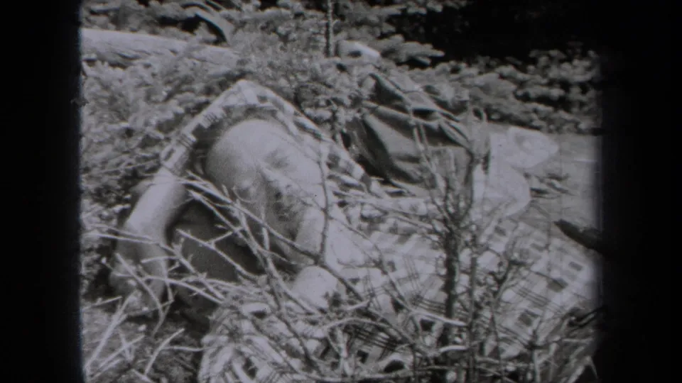 A man appears to lie or sleep among brush and a plaid blanket in 1938 New York 8mm wilderness footage.