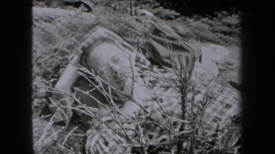 The 1938 New York home-movie frame shows a man lying low in brush, partly covered or surrounded by plaid fabric.