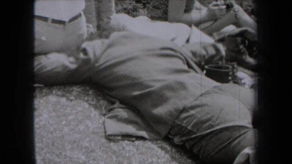A suited man lies on his side on rocky ground in 1938 New York 8mm footage, with partial figures standing nearby.