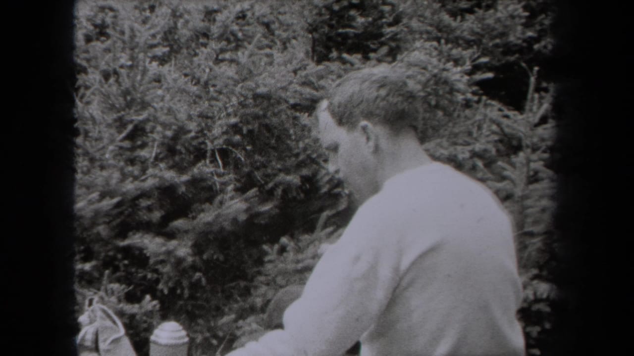 In this 1938 New York 8mm frame, a man in a light shirt sits outdoors among evergreen trees with a small container nearby.